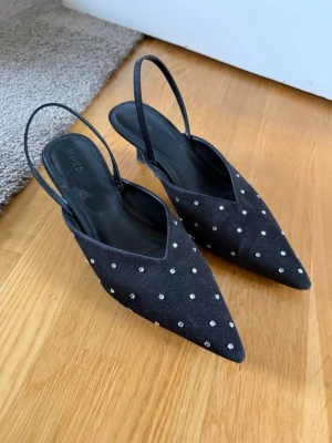 Svarta pumps med strass och slingback - Säljer ett par svarta pumps från NA-KD med spetsig tå, låg klack och slingback-rem. Skorna är dekorerade med små strassdetaljer över hela ovandelen och har en elegant siluett. Materialet är textil med en mjuk finish.