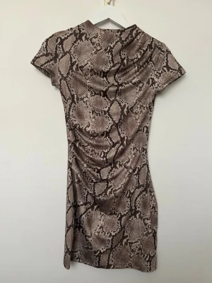 Kort klänning med ormmönster - H&M - Kort klänning med brunt och beige ormmönster. Klänningen har korta ärmar och figurnära passform, perfekt för dig som vill sticka ut. Materialet känns mjukt och stretchigt. Inköpt på H&M. Använd en gång och i mycket fint skick. Storlek S. 
