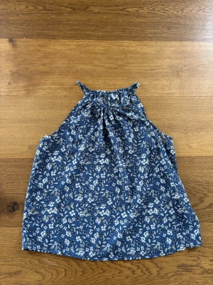 Blommig blå ärmlös topp från ONLY - Säljer en söt blå ärmlös topp från ONLY med vitt blommönster och hög hals med rynkdetalj. Perfekt för varma dagar och enkel att styla med jeans eller kjol. Luftigt material som känns skönt mot huden.