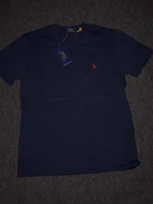 Marinblå t-shirt från Polo Ralph Lauren - Klassisk marinblå t-shirt från Polo Ralph Lauren med rund halsringning och den ikoniska röda logobrodyrn på bröstet. Tillverkad i mjuk bomull för en skön känsla och enkel stil. Perfekt till jeans eller shorts för en clean look.