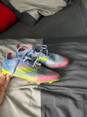 Adidas X Speedflow färgglada fotbollsskor - Säljer ett par Adidas X Speedflow fotbollsskor med snörning. Skorna har en färgstark design i blått, rosa och neongult med klassiska Adidas-ränder på sidan. Yttermaterialet är syntetiskt och sulan har fasta dobbar för spel på gräs.