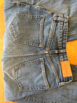 Blå raka jeans från Monki - Ett par klassiska blå jeans från Monki med raka ben och hög midja. Jeansen har fem fickor, synliga sömmar och en Monki-lapp bak i midjan. Tillverkade i slitstark denim med en avslappnad passform som passar perfekt till vardagslooken.