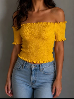 Gul offshoulder topp med volang strl  XL - Trendig gul offshoulder topp med smockad struktur och volangdetaljer vid ärmslut, halsringning och nederkant. Kortärmad och figurnära passform som ger en fräsch och somrig look. Perfekt att styla med höga jeans eller kjol.