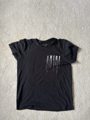 Svart AMIRI t-shirt med tryck - Svart t-shirt från AMIRI med stort tryck på ryggen och mindre tryck på bröstet. Trycket består av AMIRI-loggan med färgstänk i vitt, blått och orange som rinner ner. Klassisk passform och rund hals. Perfekt för dig som gillar streetwear och statement-plagg.