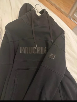 Svart hoodie från Knuckle - Säljer en svart hoodie från Knuckle med stor huva och dragsko. Hoodien har ett broderat Knuckle-tryck på bröstet och en patch på ärmen. Tillverkad i mjukt material, perfekt för en avslappnad streetwear-look.