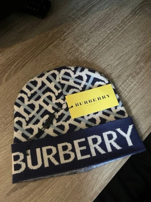  Burberry mössa - Säljer en snygg blåvit mössa från Burberry med stort logotyp-tryck framtill och klassiskt monogrammönster. Mössan är stickad i mjukt material och har en bred uppvikt kant. Perfekt accessoar för dig som vill ha en trendig och exklusiv look.