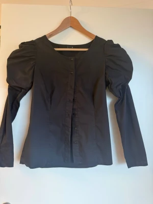 Svart blus med puffärmar - Stilren svart blus med markerade puffärmar och knappar framtill. Blusen har rund halsringning och är figursydd för en snygg siluett. Tillverkad i ett slätt tyg som ger en klassisk look. Perfekt för dig som gillar trendiga detaljer och enkel design.