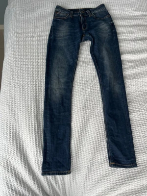 Mörkblå skinny jeans från Nudie - Säljer ett par mörkblå skinny jeans från Nudie med snygga slitningar och kontrastsömmar. Klassisk femficksmodell med läderpatch bak i midjan och broderade bakfickor. Perfekta för dig som gillar en tight passform och streetig look.
