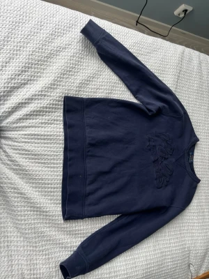 Mörkblå sweatshirt från Polo Ralph Lauren - Snygg mörkblå sweatshirt från Polo Ralph Lauren med broderad logga på bröstet. Tröjan har rund halsringning och ribbade muddar vid ärmslut och nederkant. Perfekt för en avslappnad och stilren look.