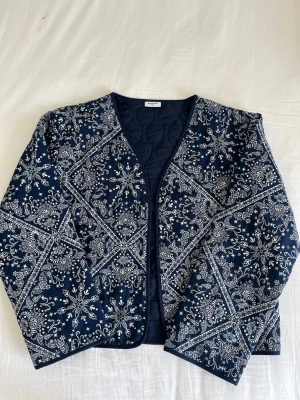 Mörkblå quiltad cardigan från Kappahl - Snygg mörkblå quiltad cardiganjacka från Kappahl med vitt paisleymönster över hela plagget. Jackan har rund halsringning, raka ärmar och är lätt vadderad för extra komfort. Perfekt för dig som vill ha en unik och trendig look. Storlek 170. Aldrig använt.