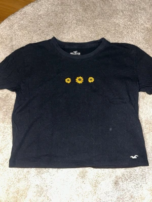 Svart croppad t-shirt Hollister - Svart croppad t-shirt från Hollister med tre broderade gula blommor på bröstet och liten logga nere vid fållen. Tillverkad i 100% bomull och har en klassisk rund halsringning och korta ärmar. Perfekt för en chill och trendig look.