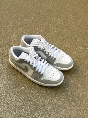 Nike Air Jordan 1 Low grå/vit - Snygga Nike Air Jordan 1 Low sneakers i grått och vitt. Skorna har klassisk låg siluett, perforerad tåbox och vita skosnören. Tillverkade i läder med ikoniska swoosh-loggan på sidan. Perfekta för dig som gillar stilrena och tidlösa sneakers.