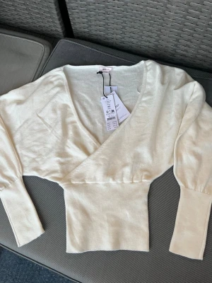 Krämvit omlottstickad topp H&M - Säljer en krämvit stickad topp från Gina Tricot med omlott-design och bred ribbad midja. Tröjan har djup v-ringning och långa ärmar med breda muddar. Perfekt för en trendig och avslappnad look. Materialet är mjukt och stretchigt. 