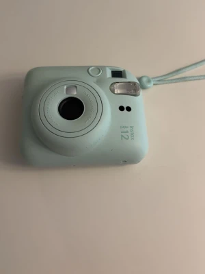 Instax Mini 12 kamera - Hej! Säljer en ljusblå Fujifilm Instax Mini 12 direktfilmskamera. Kameran har rundad form, inbyggd blixt och enkel knapp för att ta bilder. Tillverkad i plast och har en matchande handledsrem. Perfekt för att ta roliga polaroidbilder med vänner eller för att fånga mysiga ögonblick. Hör av dig vid mibsta fundering! 