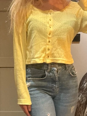 Gul cropped cardigan med knappar - Snygg gul cardigan med långa ärmar och knäppning framtill. Croppad modell med diskret mönster i tyget, perfekt att styla med höga jeans. Mjuk och lätt i materialet, passar för vår och sommar. Skulle uppskatta till strlk s-m