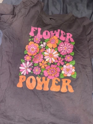 Grå Flower Power t-shirt - Cool grå t-shirt med färgglatt tryck av blommor och texten 'Flower Power' i retrostil. Blommorna är i rosa, orange och vitt med gröna blad. Perfekt för dig som gillar vintage vibes och statement-plagg.