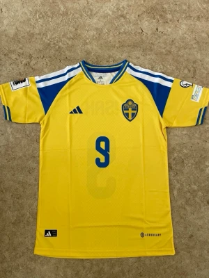 Sverige matchtröja Isak nr 9 Adidas - Gul och blå matchtröja från Adidas för svenska landslaget med Isak och nummer 9 på ryggen. Tröjan har korta ärmar, klubbmärke på bröstet och AEROREADY-material som andas. Perfekt för fotbollsträning eller att bära på läktaren.