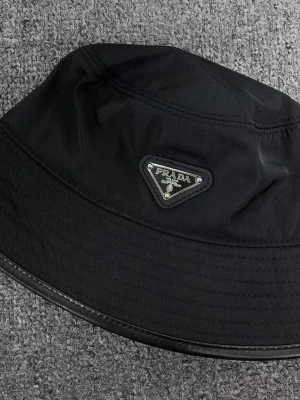 Svart bucket hat från Prada - Snygg svart bucket hat från Prada med den ikoniska triangulära logotypen framtill. Hatten är tillverkad i nylon med bomullsfoder, vilket gör den både lätt och bekväm. Perfekt accessoar för en trendig streetwear-look.