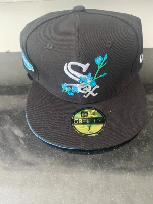 Svart New Era 59Fifty White Sox keps - Snygg svart 59Fifty keps från New Era med White Sox-logga framtill och blåa blommor broderade runt loggan. Klassisk flat brim och broderade detaljer på sidan. Tillverkad i bomull och polyester, perfekt för dig som vill sticka ut med en unik keps.