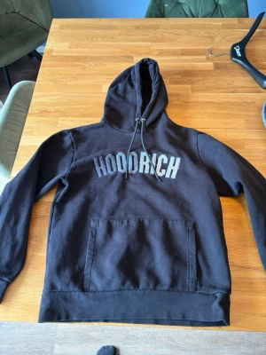Svart hoodie från Hoodrich - Säljer en svart hoodie från Hoodrich med stor broderad logga på bröstet. Hoodien har huva med dragsko och en stor magficka. Perfekt för dig som gillar streetwear och vill ha en chill och snygg look.