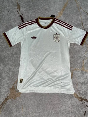 Spanien borta  - Säljer en vit Spanien fotbollströja från Adidas med vinröda detaljer och tre ränder på axlarna. Tröjan har korta ärmar, rund halsringning och broderad landslagssköld på bröstet. Tillverkad i lätt och ventilerande material, perfekt för träning eller match. M för en tajtare passform, passar även S för mer normal passform 