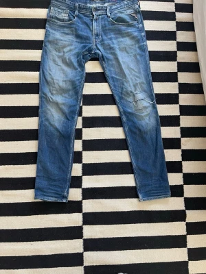 Blå slitna slim fit jeans från replay  - Säljer ett par blå replay  med slitningar och tvättade detaljer på låren och knäet. Jeansen har fem fickor, normal midja och klassisk dragkedja med knapp. Perfekta för en avslappnad och trendig look.