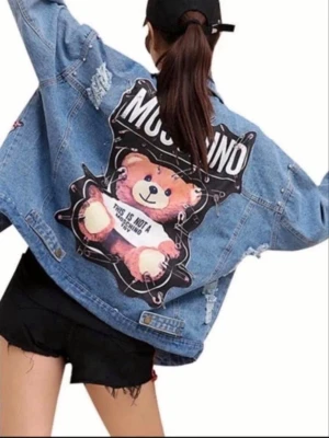 Moschino jeansjacka med björntryck - Oversized ljusblå jeansjacka från Moschino med stort björntryck och text på ryggen. Jackan har slitna detaljer, pärlor och patchar, samt tryckta loggor. Perfekt för dig som vill sticka ut och älskar statement-plagg.