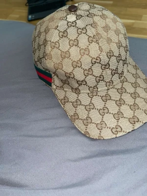 Beige keps från Gucci med GG-mönster - Säljer en beige keps från Gucci med det klassiska GG-monogrammet över hela kepsen. På sidan finns en grön och röd rand som ger extra lyxig känsla. Kepsen har böjd skärm och är tillverkad i canvasmaterial. Perfekt för dig som vill ha en ikonisk accessoar.