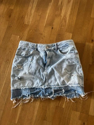 Lågmidjad jeanskjol - Trendig ljusblå jeanskjol med slitna detaljer och fransig, rå kant längst ner. Kjolen har klassisk femficksdesign och bälteshällor. Perfekt för dig som gillar en avslappnad och edgy stil.