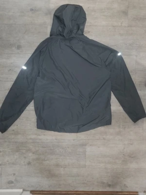 Nike grå vindjacka med huva - Snygg grå vindjacka från Nike med huva och reflekterande detaljer på ärmarna. Jackan har dragkedja framtill och är tillverkad i lätt, vindtätt material. Perfekt för löpning eller när du vill ha en sportig look.