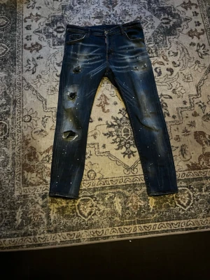 Dsquared2 jenas blå - Säljer ett par mörkblå jeans från Dsquared2 med slitningar och hål på benen samt vita färgstänk. Jeansen har klassisk femficksmodell, knappgylf och kontrastsömmar. Perfekt för dig som gillar en edgy och trendig stil.