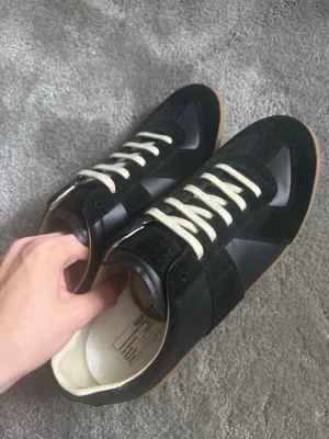Maison Margiela svarta sneakers - Snygga svarta sneakers från Maison Margiela med beige sula och vita skosnören. Skorna har en mix av mocka och läder på ovandelen och klassisk låg siluett. Perfekta för dig som gillar stilrena och tidlösa sneakers med exklusiv känsla.
