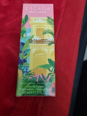 Escada Brisa Cubana Eau de Toilette - Fräsch och tropisk Eau de Toilette från Escada, Brisa Cubana. Flaskan är elegant med silverfärgad kork och rosa tonat glas. Förpackningen har färgglada illustrationer med tropiska frukter och växter, vilket ger en somrig och livlig känsla.
