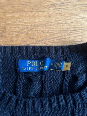 Mörkblå kabelstickad tröja Polo Ralph Lauren - Kabelstickad mörkblå tröja från Polo Ralph Lauren med rund halsringning och röd broderad logga på bröstet. Tröjan har klassisk passform och ribbade muddar. Perfekt för dig som gillar tidlös och stilren design.