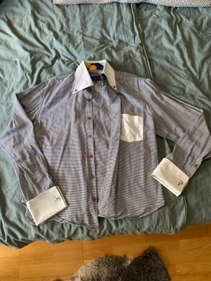 Rutig skjorta från Evisu Deluxe  - Unik rutig skjorta från Evisu Deluxe med kontrasterande vita manschetter, krage och bröstficka. Skjortan har röda knappar och ett färgglatt mönster på insidan av kragen och handled. Perfekt för dig som gillar detaljer och vill sticka ut.