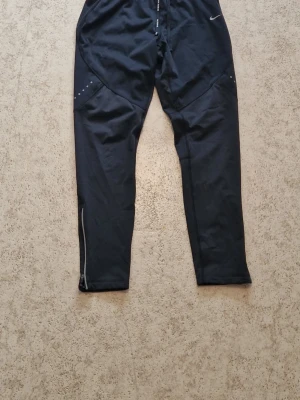 Svarta träningsleggings från Nike - Säljer ett par svarta träningsleggings från Nike med reflekterande detaljer och smal passform. De har dragsko i midjan och små vita loggor på benet. Perfekta för löpning eller gymmet, tillverkade i stretchigt funktionsmaterial.
