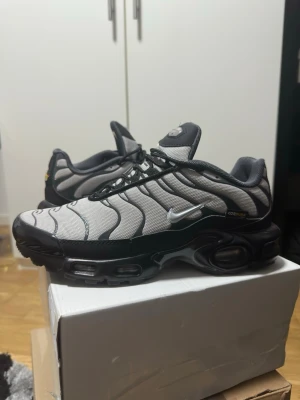 Nike Air Max Plus TN Cordura  - Nike Air Max Plus TN sneakers i grått och svart med vita detaljer. Skorna har den ikoniska vågiga designen på ovandelen, synlig Air-dämpning i sulan och små gula detaljer vid hälen. Tillverkade i mesh och syntetmaterial med klassisk Swoosh-logga på sidan.
