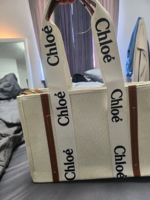 Chloé Woody beige handväska - Snygg beige handväska från Chloé med tydliga logoband i vitt och svart text. Väskan har rektangulär form, detaljer i brunt läder och breda handtag i canvas. Perfekt för dig som gillar trendiga accessoarer och vill sticka ut med en ikonisk design. Ej äkta