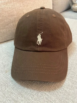 Brun keps från Polo Ralph Lauren - Säljer en brun keps från Polo Ralph Lauren med klassisk böjd skärm och broderad logga framtill. Baksidan har justerbart spänne och broderad POLO-text. Tillverkad i bomullstyg som känns både slitstarkt och bekvämt.