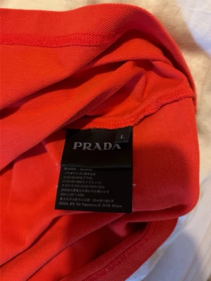 Röd pikétröja från Prada - Snygg röd pikétröja från Prada med klassisk krage och knappar framtill. Tröjan har en sportig look med ribbade detaljer på kragen och är tillverkad i mjuk bomull som känns skön mot huden. Perfekt för dig som vill ha en stilren och exklusiv skjorta.