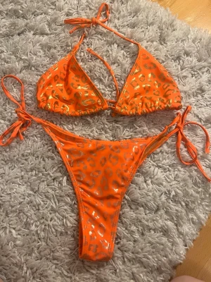 Orange bikini med leopardmönster - Snygg orange bikini med glittrigt leopardmönster i guld. Klassisk trekantsmodell på överdelen och knyt i både sidor och nacke för perfekt passform. Perfekt för sommarens strandhäng eller poolparty.