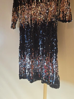 Glittrig paljettklänning i svart och guld - Säljer en kort glittrig klänning täckt av paljetter i svart, silver och guld. Klänningen har halvlånga ärmar och en rak passform som ger en cool och festlig look. Perfekt för dig som vill sticka ut på nästa fest! ✨