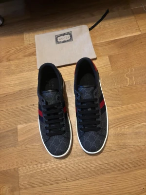 Gucci sneakers med monogram och röd detalj - Snygga sneakers från Gucci i mörkblått tyg med klassiskt GG-monogram över hela skon. Skorna har svarta skosnören, vit sula och de ikoniska röd-blå ränderna på sidan. Perfekta för dig som vill ha en exklusiv och trendig look.