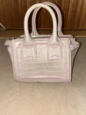 Ljusbeige croco handväska med rosa sömmar - Säljer en liten handväska i ljusbeige med croco-mönster och rosa kontrastsömmar. Väskan har två korta handtag och dragkedja upptill. Perfekt för dig som gillar trendiga detaljer och en unik look. Materialet är konstläder.
