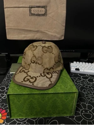 Beige keps med GG-mönster från Gucci - Säljer en beige keps från Gucci med det klassiska GG-monogrammet i brunt över hela kepsen. Kepsen har böjd skärm och är tillverkad i canvasmaterial med bruna detaljer. Perfekt för dig som vill ha en stilren och lyxig accessoar.