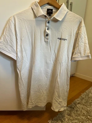 Vit pikétröja från Armani Exchange - Klassisk vit pikétröja från Armani Exchange med krage och knappar framtill. Diskret logga på bröstet och korta ärmar. Perfekt till jeans eller shorts för en clean och stilren look.