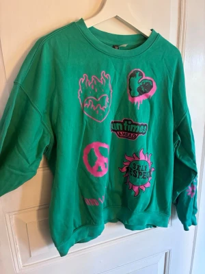 Grön sweatshirt med tryck från H&M - Cool grön sweatshirt från H&M med flera färgglada tryck i rosa och svart, bland annat hjärta, eld, peace-märke och texten 'SELF RESPECT' och 'fun times ahead'. Oversized passform och rund halsringning. Perfekt för dig som gillar statement-plagg och streetwear vibes.
