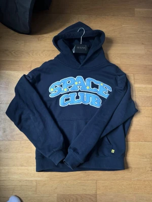 Broken Planet Hoodie - Snygg mörkblå hoodie från Broken Planet med stor tryckt text 'SPACE CLUB' i ljusblått och vitt med gula stjärnor på bröstet. Klassisk huva och magficka framtill. Perfekt för dig som gillar streetwear och vill sticka ut med coolt tryck.