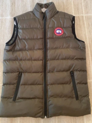 Olivgrön dunväst från Canada Goose - Snygg olivgrön dunväst från Canada Goose med hög krage och dragkedja framtill. Västen har två fickor med dragkedja och den klassiska röda logotypen på bröstet. Perfekt för lager-på-lager och håller dig varm under kyliga dagar.