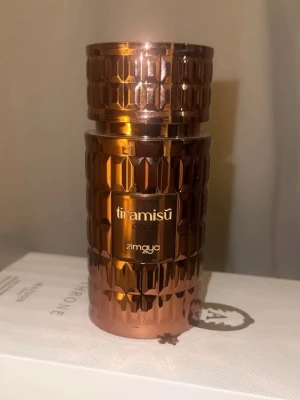 Tiramisu Coco parfym från Zimaya - Lyxig parfymflaska i kopparfärgat metall med geometriskt mönster. Märkt med 'tiramisu coco' och 'zimaya'. Flaskan har en modern, elegant design som sticker ut på parfymhyllan.